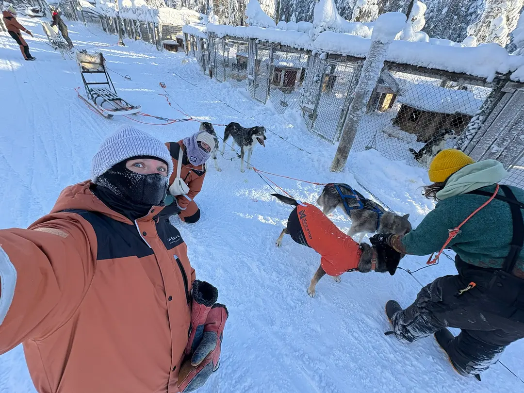 Group selfie of bundled-up guests with huskies at a snowy husky farm