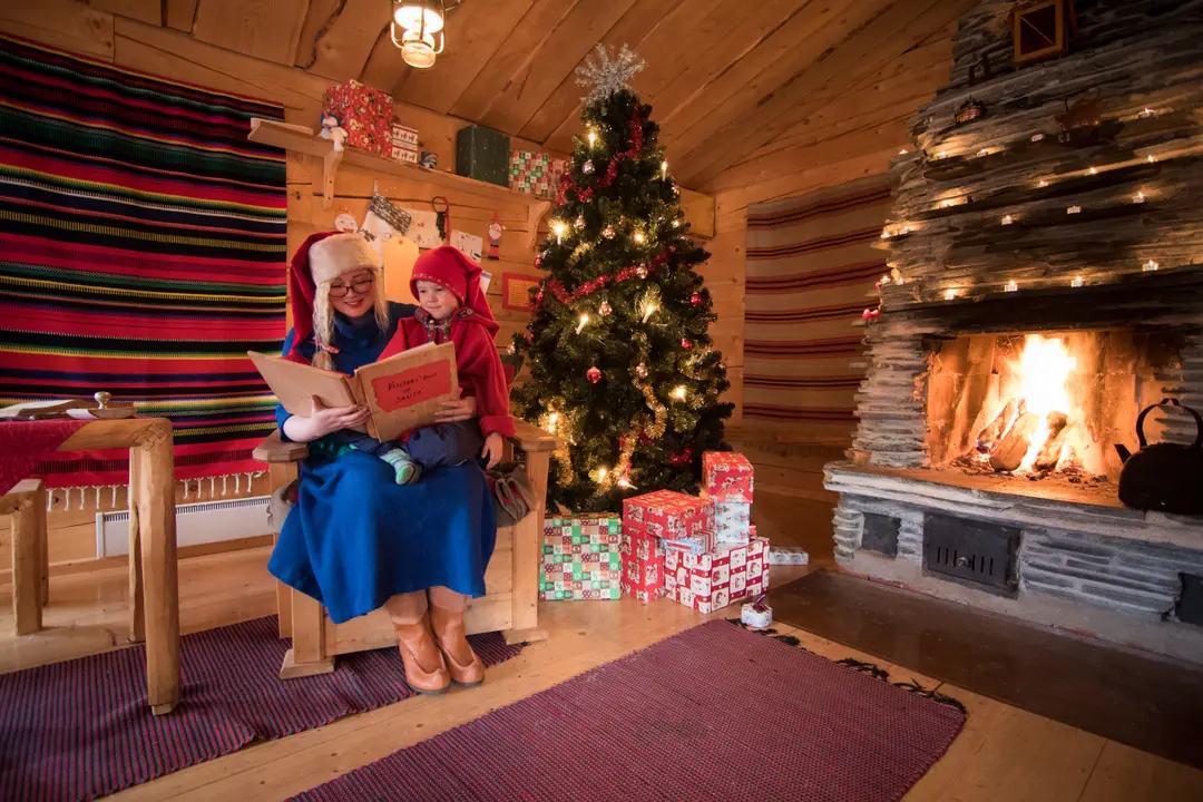 Mrs Claus reading a storybook to a child dressed as an elf beside a Christmas tree and glowing fireplace