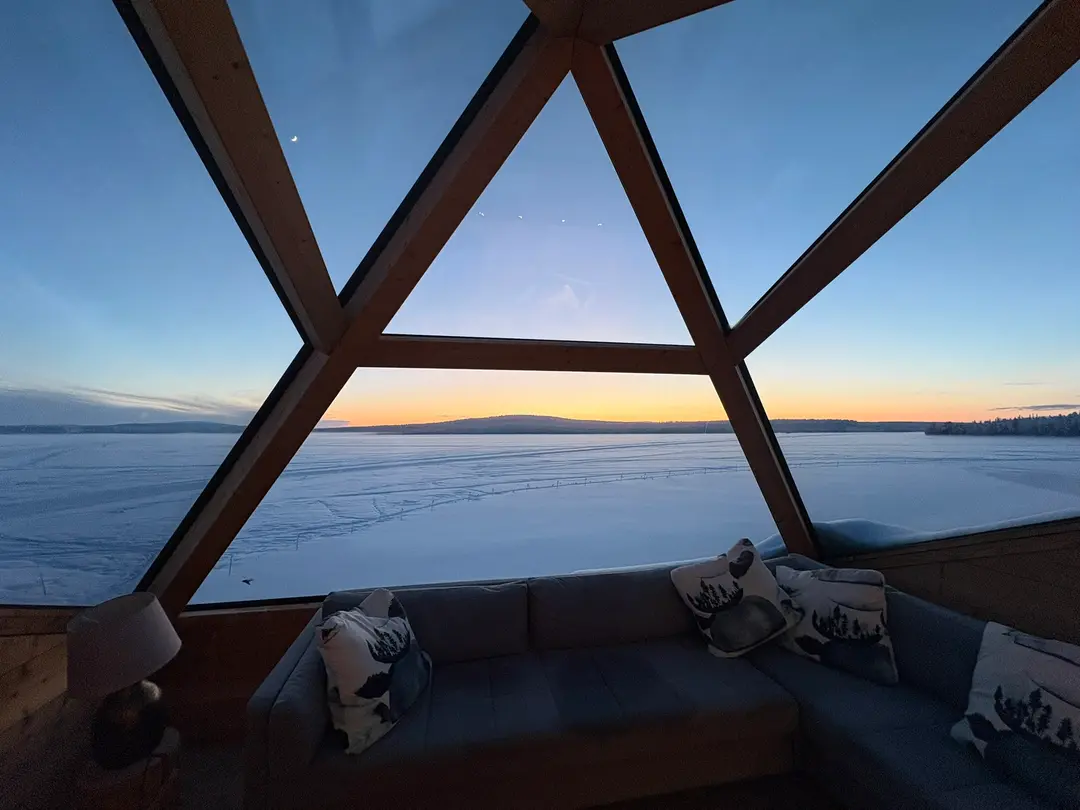 Cosy glass-walled cabin interior looking out over a frozen lake at sunset